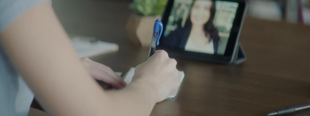 A woman writing in a small notebook while watching a video on a tablet