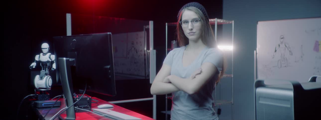 A young woman stands at a computer while typing before crossing her arms and looking confident