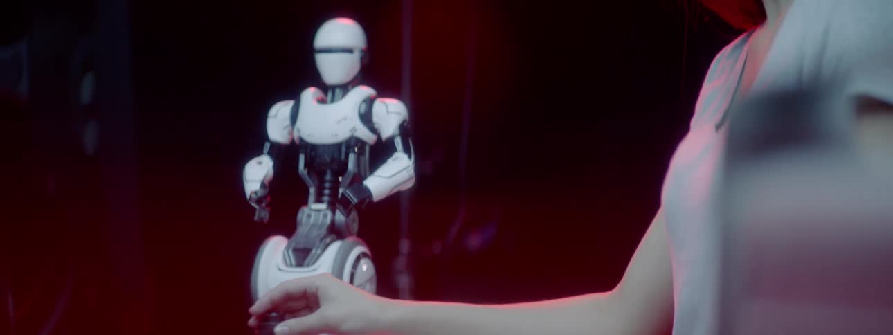 A woman sits at a computer and adjusts a controller which moves a small white robot next to her