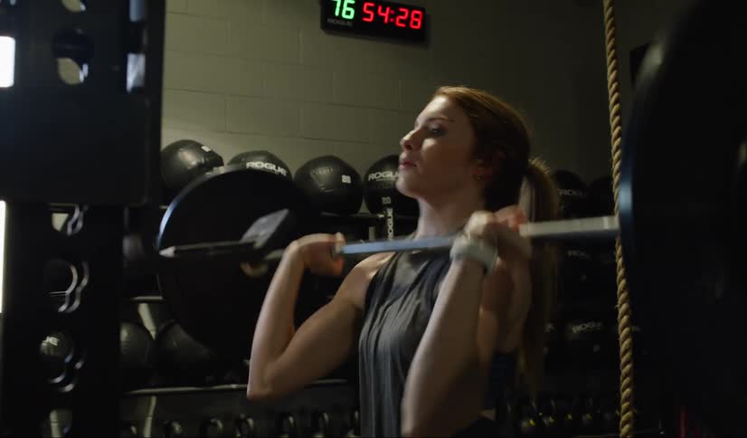 A woman in a gym does overhead presses with a barbell and weights