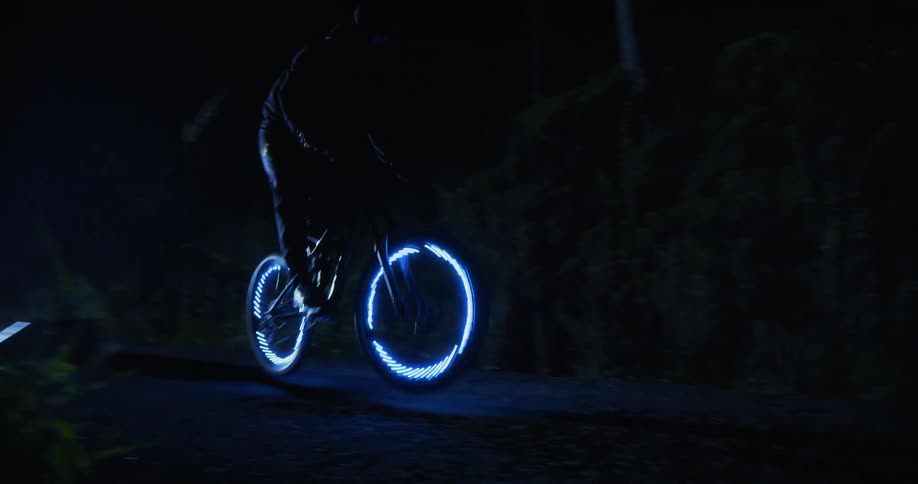 A person rides a bike with blue lights affixed to the wheels through a darkened outdoor area