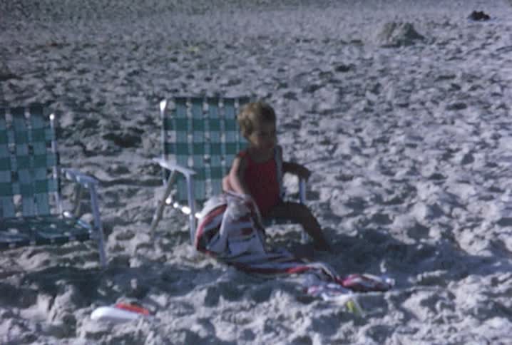 A small boy plays in the sand on a beach while a little girl sits in a chair watching