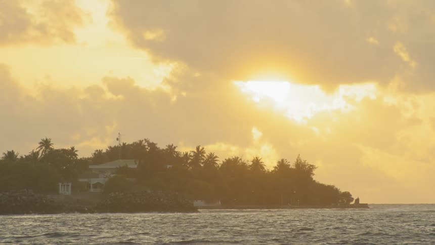 The sun peeks through the clouds over a house next to the ocean surrounded by palm trees