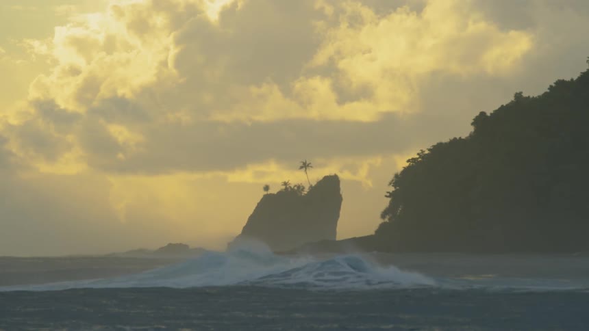 A wave crashes along the ocean surface next to large hills and coastal mountains
