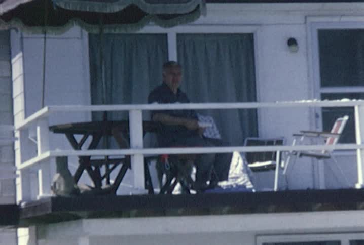 A man sits at a table under an umbrella on a balcony