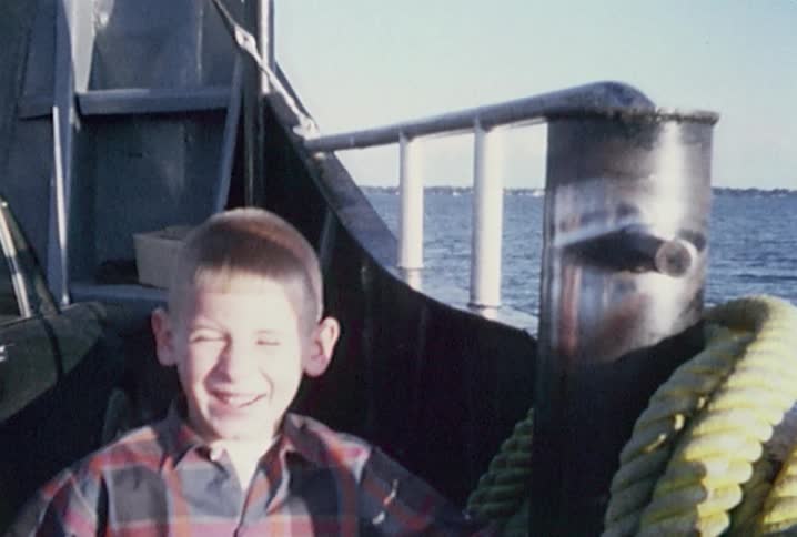 Old footage of  young boy talking and grinning at the camera while on a ferry boat crossing a body of water