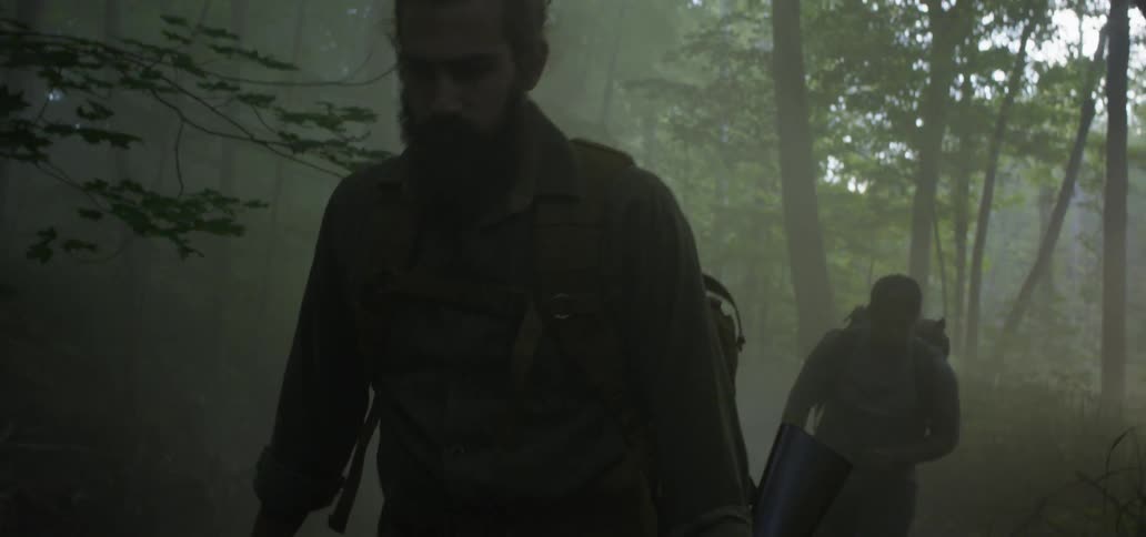 Two men are walking through the woods one carrying a rifle and the other a large knife