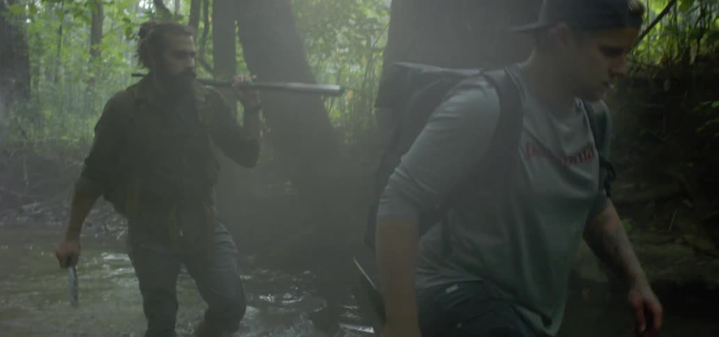 Men with backpacks walking on shallow river through forest