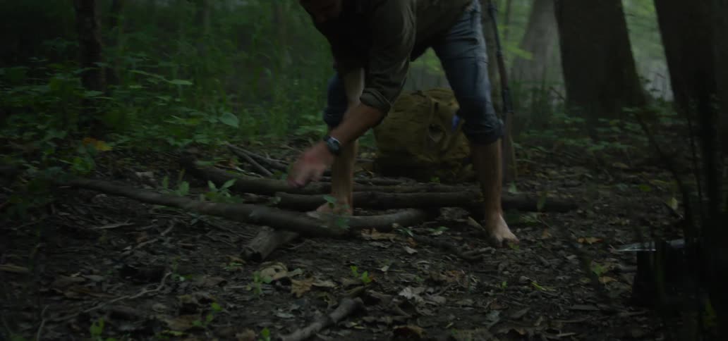 A man walks through a forest with a knife and then starts hitting a branch with it