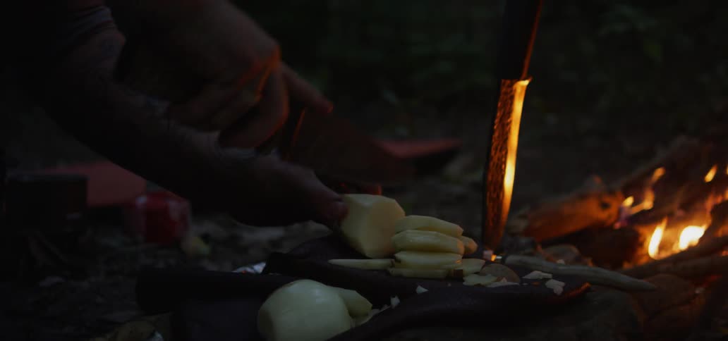 A person uses a knife to cut potato outside by a campfire