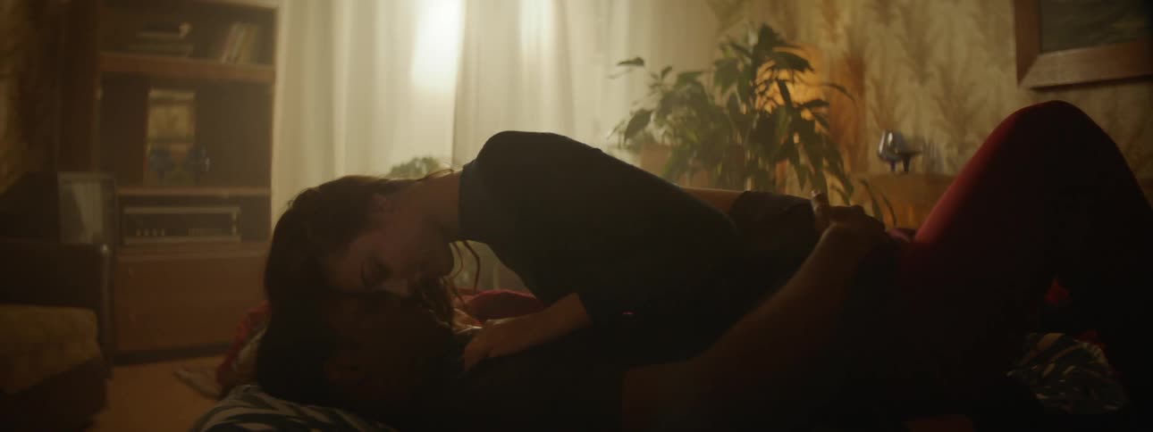 A man rolls over in bed pulling a woman on top of him as they embrace and kiss