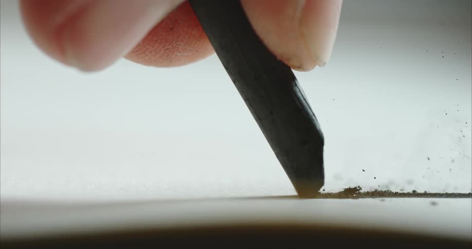 A person drags a piece of graphite across a paper and chunks of its tip break off and are ejected