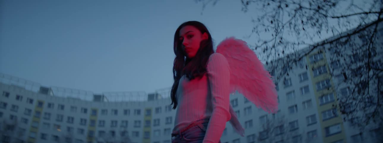 A dark haired woman with small angel wings standing under a red light in front of a large building