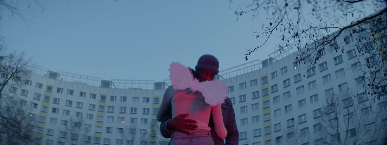 A man in a stocking cap and a woman wearing wings hug in front of a high rise building