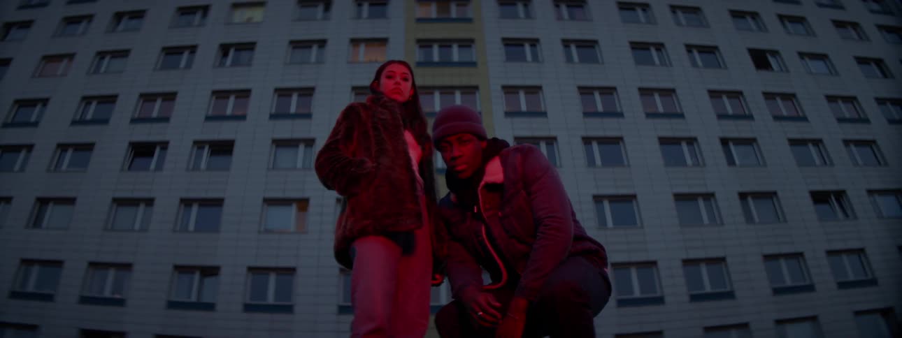 A young male and female stand before a tall apartment building as the male crouches down