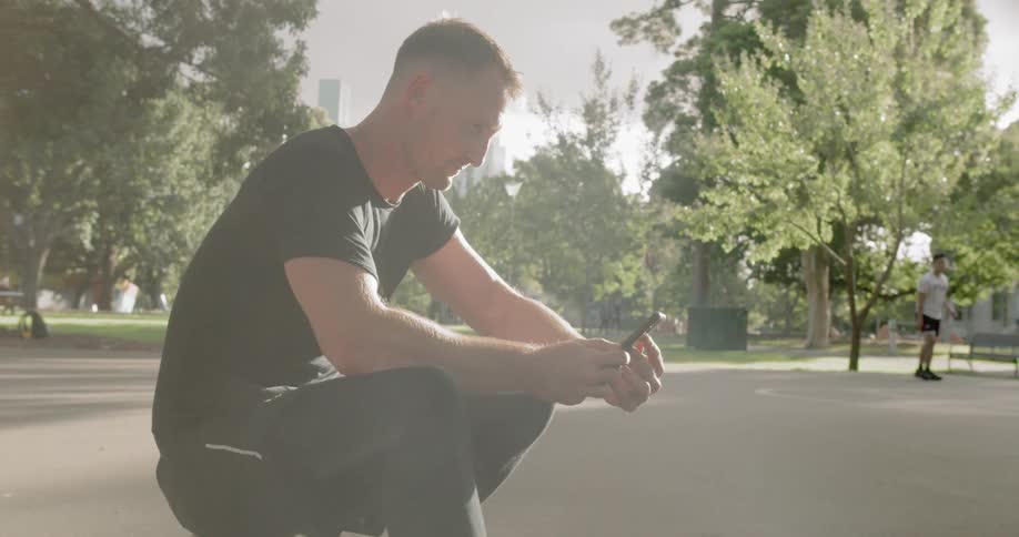 Man sits on a basketball while texting on smart phone in public park