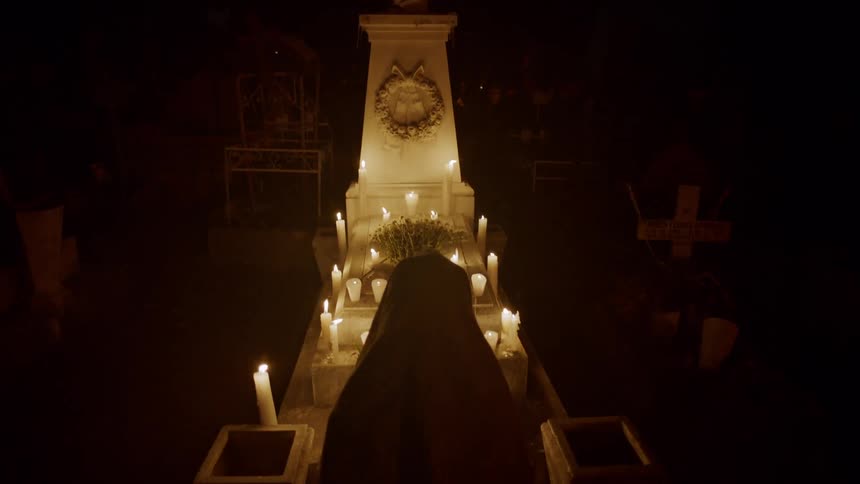 A person in a white veil kneeling in a dark room before an altar illuminated by candles