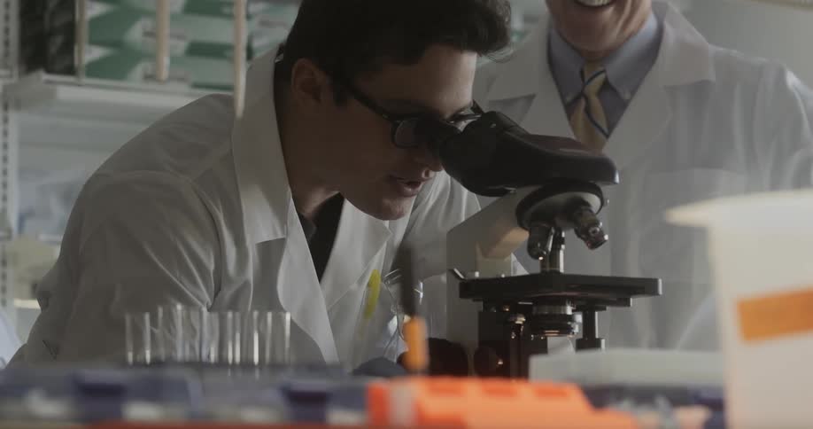 Two men in lab coats while one looks into a microscope