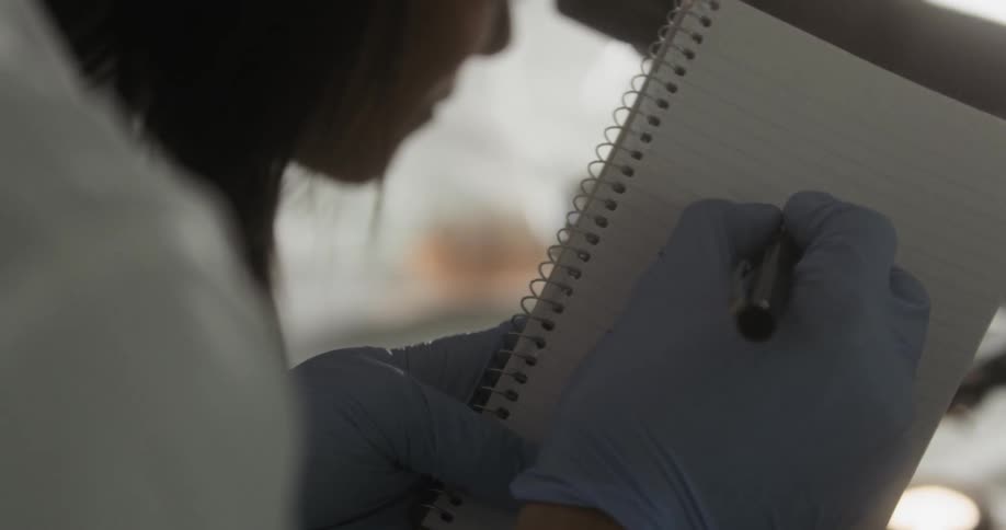 A person in a lab coat and nylon gloves writing in a notepad