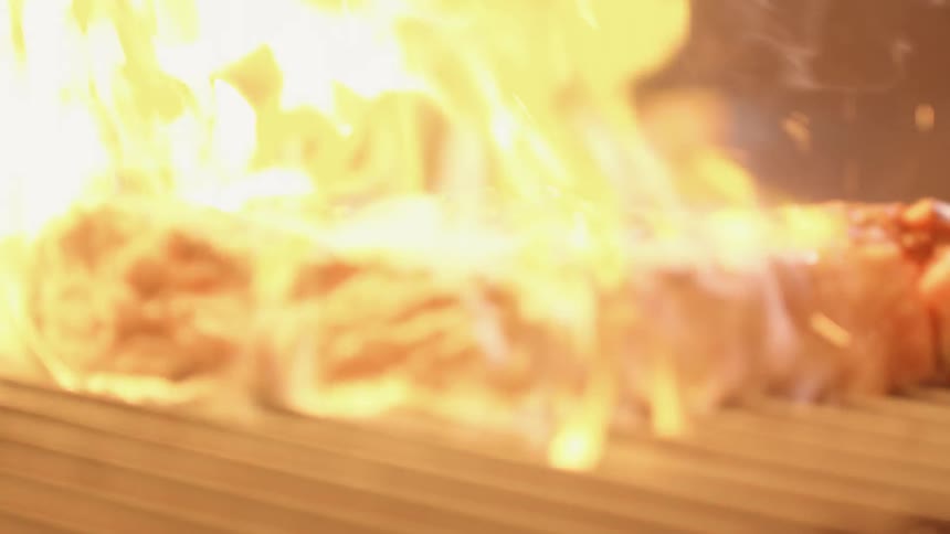 A piece of steak is being grilled over an open flame which flares up as the steak is being flipped over