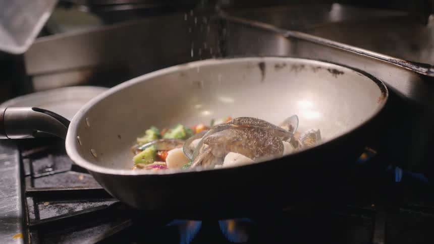 A pan of full of seafood and ingredients is heating on a gas stove as a someone sprinkles seasoning on it