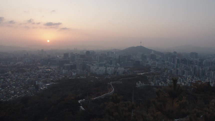 A sprawling city surrounds a tree covered cluster of hills as the sun sits low in the sky