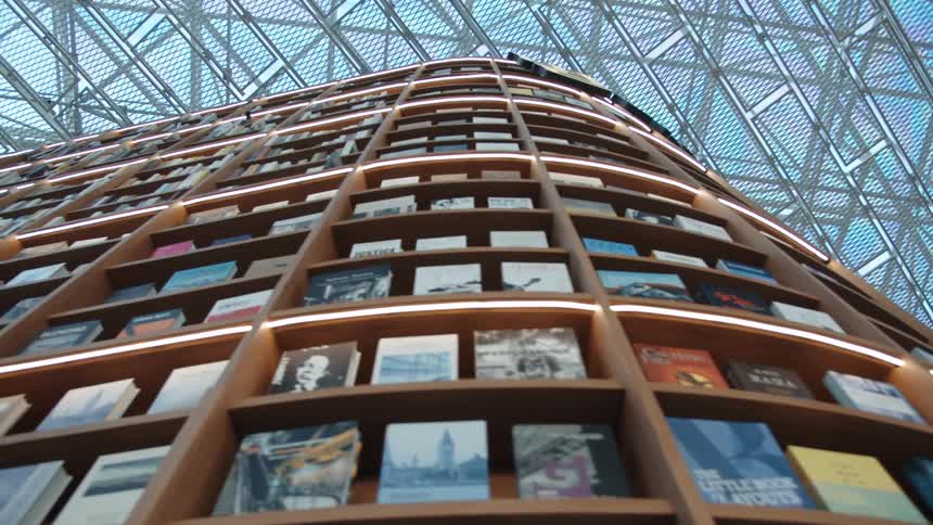 A floor to ceiling book shelf with rounded edges under a glass ceiling