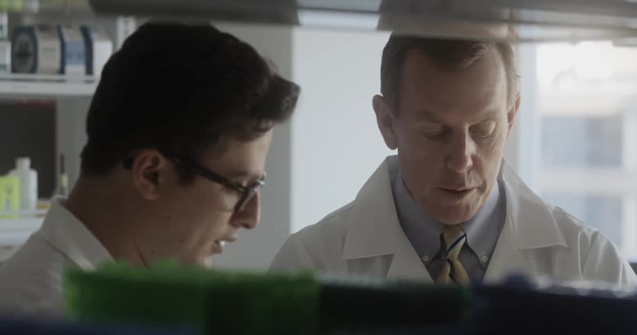 Two men in lab coats talk in front of a window in a lab