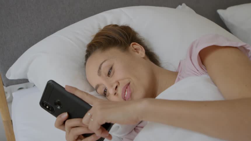 A woman smiling while using her smartphone in bed