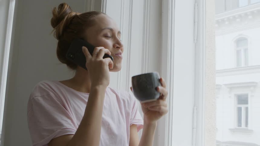 A woman talks on the phone and drinks coffee while standing next to a window