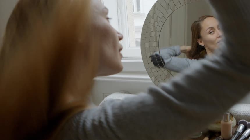A woman sits at a vanity brushing her hair