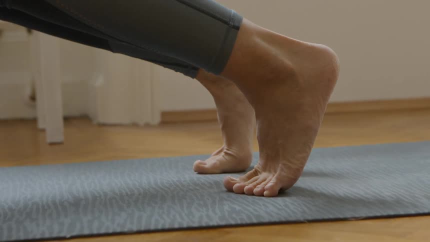 A barefoot woman goes up on her toes while doing yoga on a yoga mat