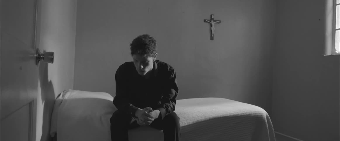 Man sits on bed on bare room with crucifix on wall
