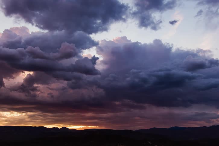 Crimson tint on dark clouds fades as sun sets over mountains on horizon