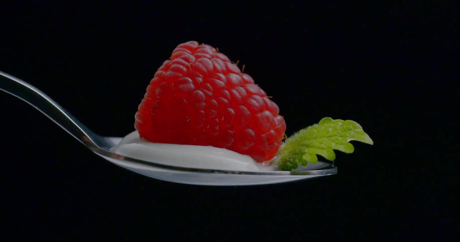 A spoon holding a dollop of yogurt a sprig of mint and a raspberry is turning around clockwise