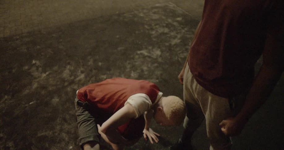 A teenager sits on the floor holding a basketball and stands up as he is surrounded by larger men