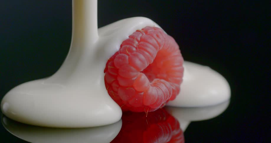 A single raspberry being covered in white cream on black background