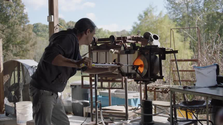 Man turning molten glass on a metal rod inside a furnace outdoors