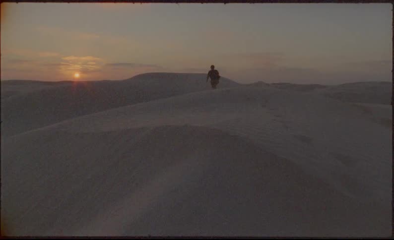 A person walks across large sand dunes towards the horizon