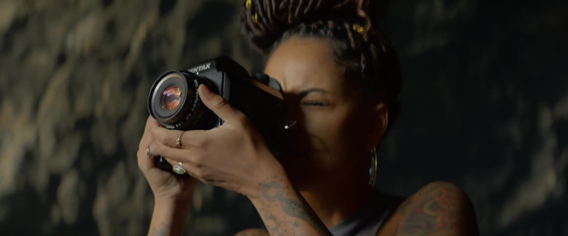 Female places the camera up to her right eye while closing her left then adjusts the zoom lens and smiles after taking a picture