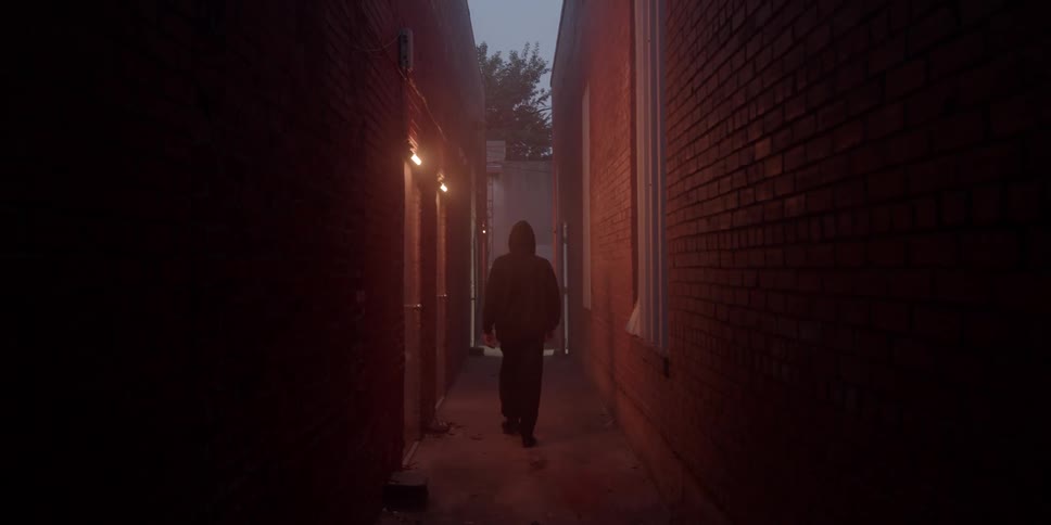 A man wearing black clothing walks down an alley at night