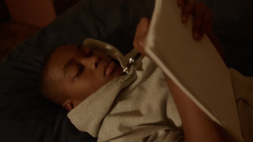 A boy wearing a hoodie writes on a notepad while laying down on a bed
