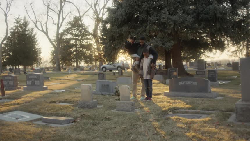 A father and two young children stand in front of a grave in a cemetery