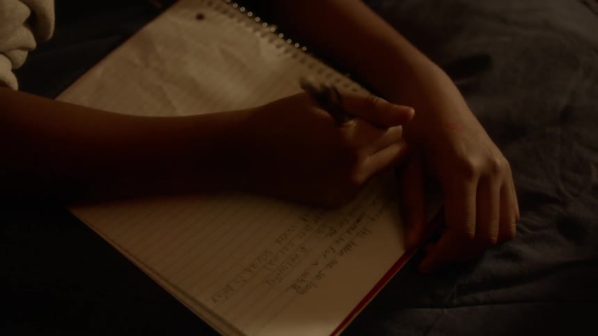 A boy writing in a notebook while lying on a bed