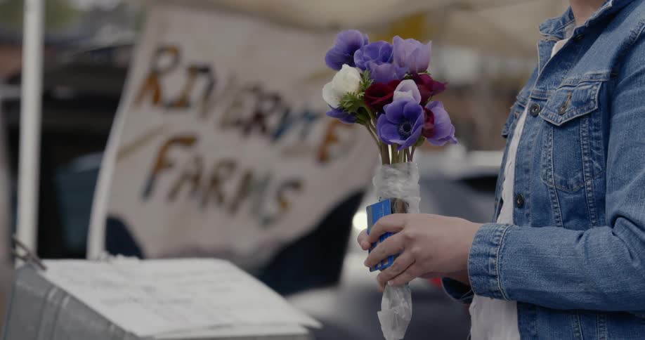 A woman wearing a denim jacket holds a small bouquet of flowers while a farm banner blows in the wind