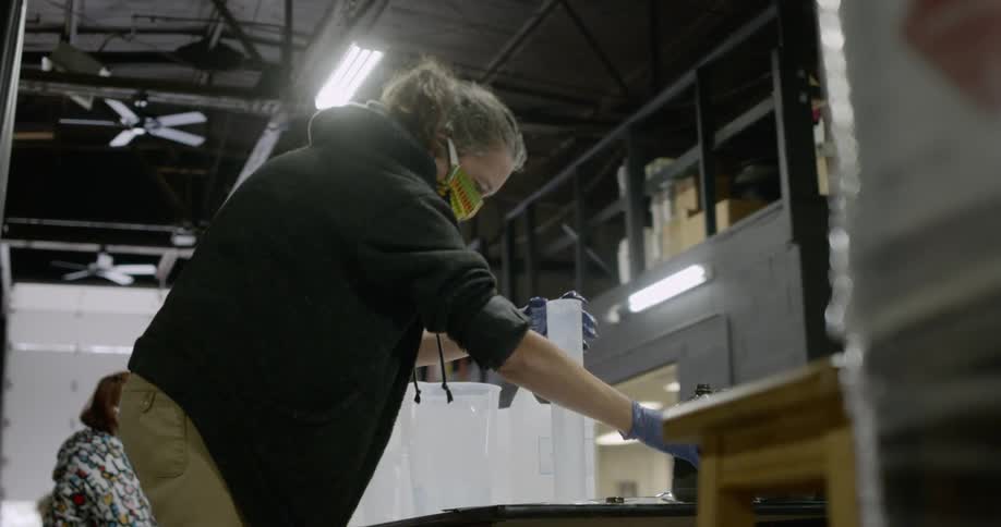 A woman in a facemask pours liquid into a graduated cylinder in a warehouse