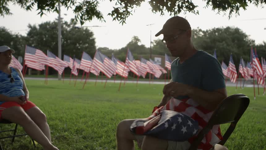 A man sits in a folding chair on a lawn and struggles with an american flag on a poll