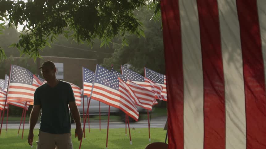 A man walks across a lawn filled with american flags and then sits in a chair
