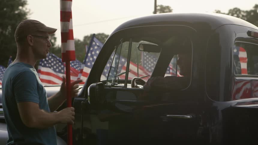 A man holding a rolled up flag shakes hands with a man in a truck