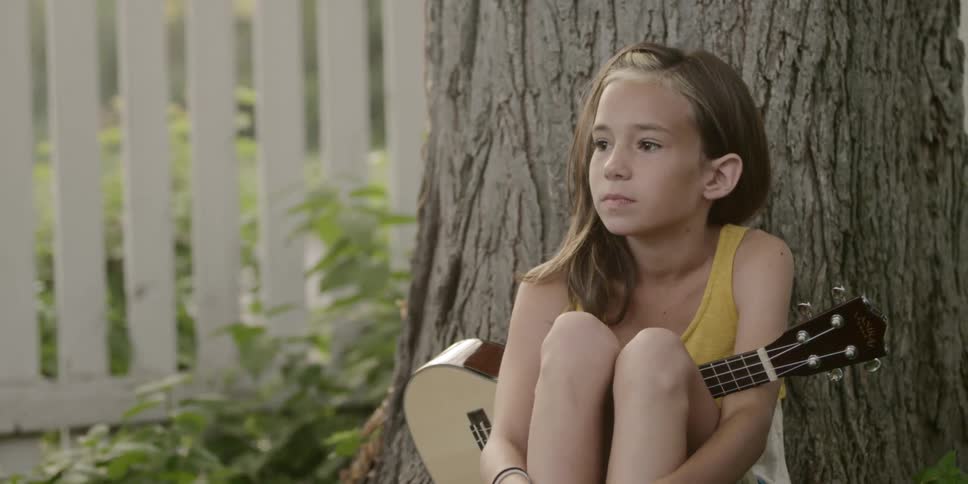 A young girl sits against a tree holding a guitar